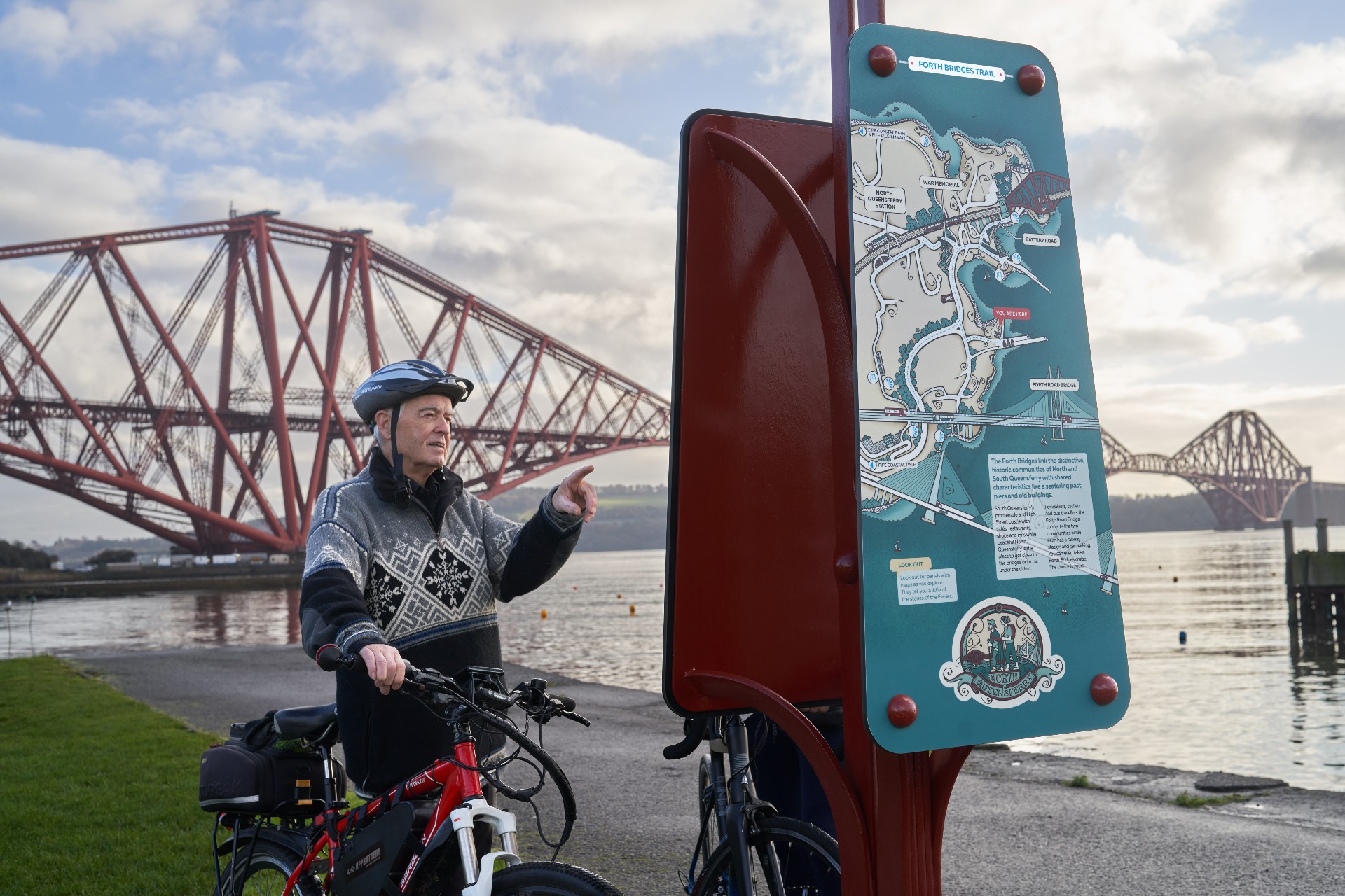 A person with a bike looking at and information board with a bridge in the background