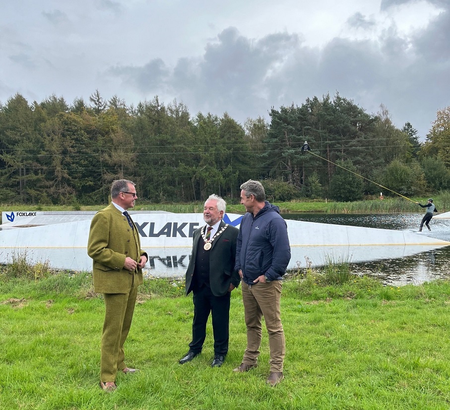 Three people standing on a grassy bank with a person doing water sports in the loch in the background