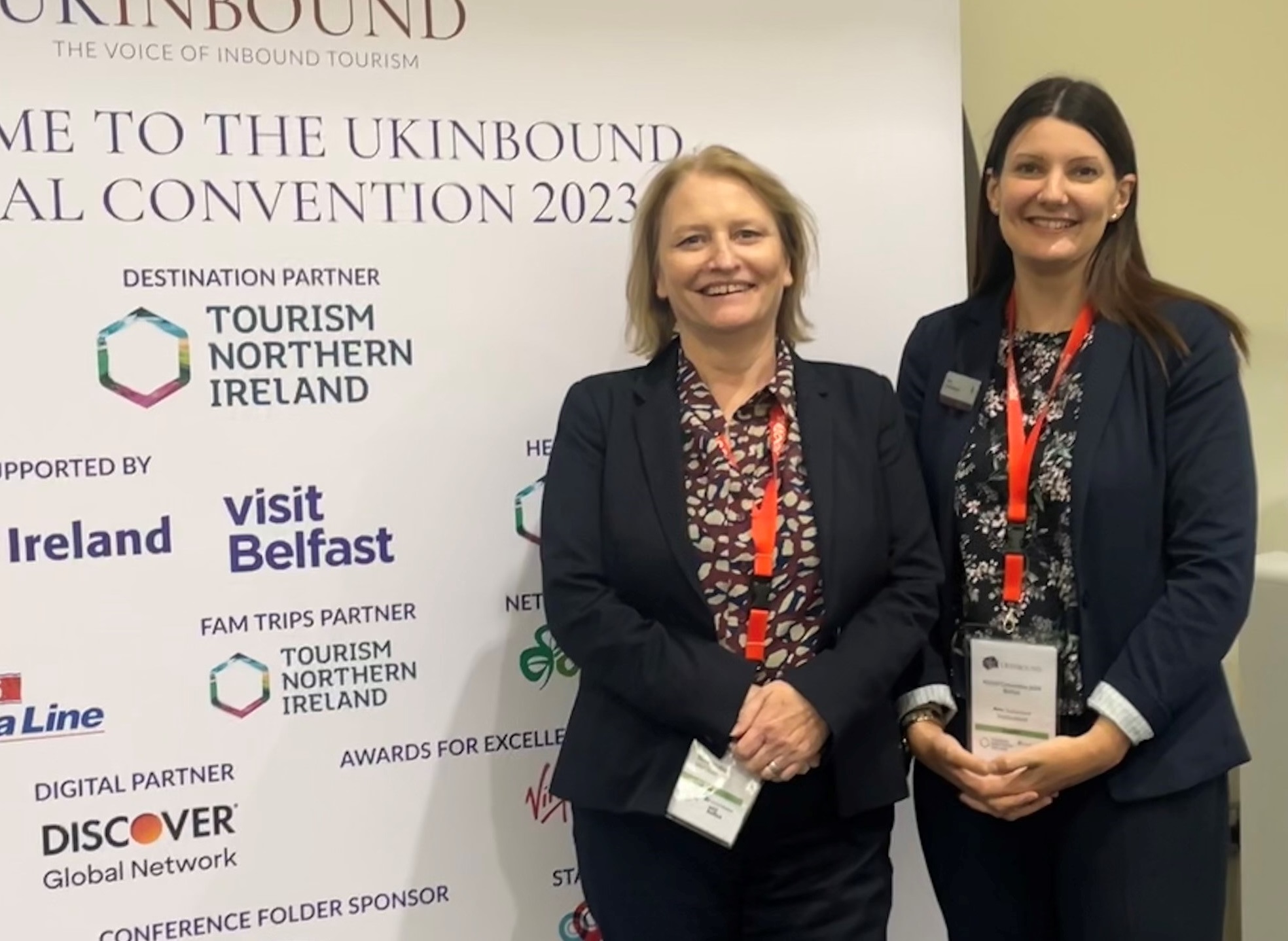 Two women pose in front of a banner of event sponsors