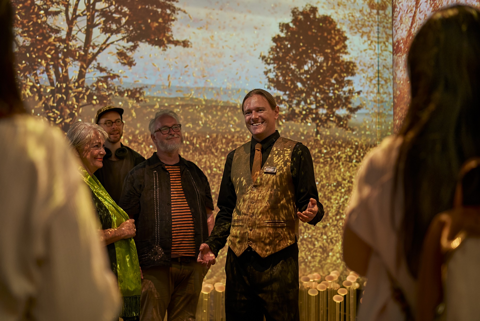 People listening to a staff member speaking on the tour with a brown and gold digital display of trees behind them.