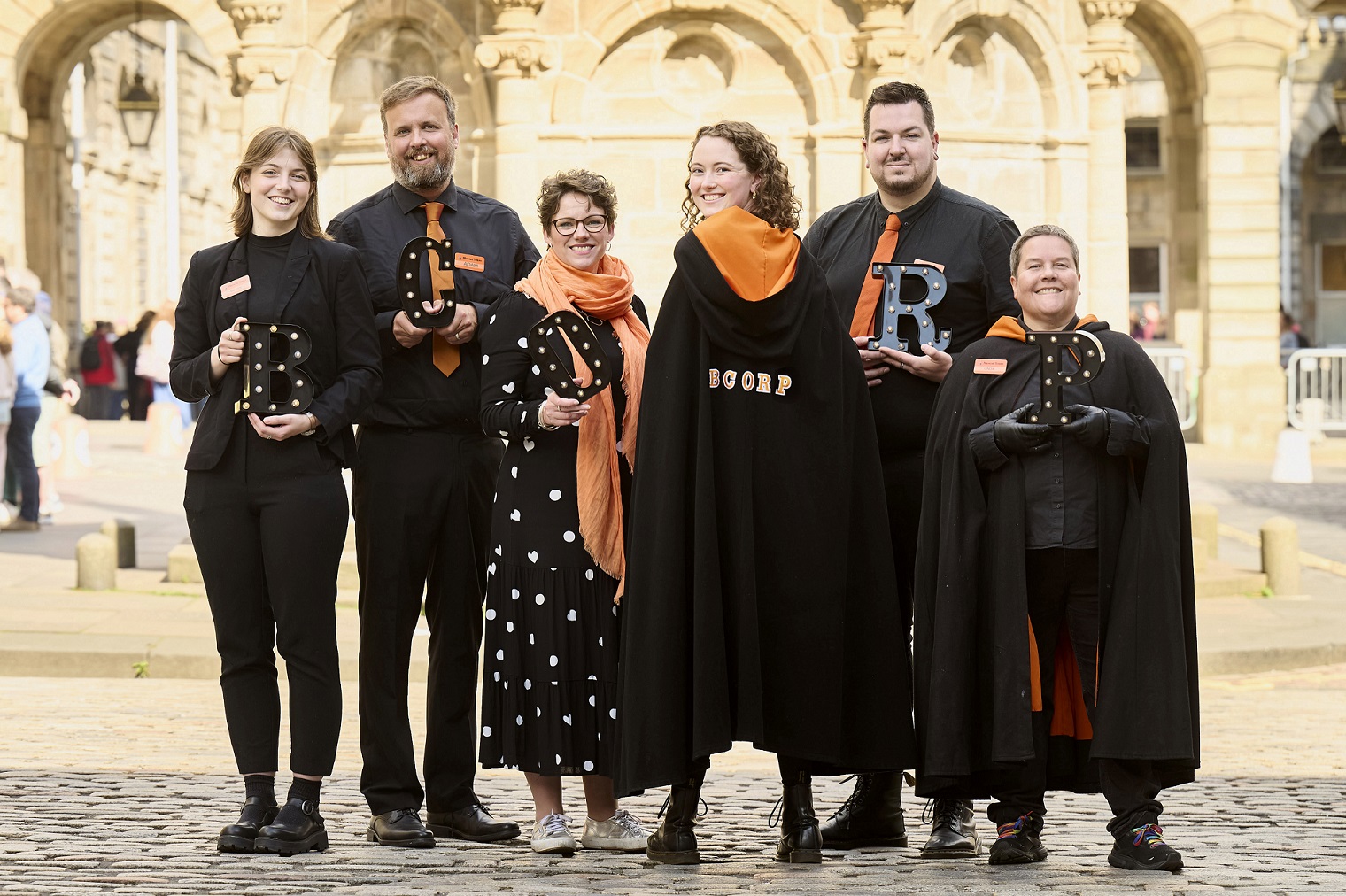 A group of people standing outside dressed in black cloaks, in front of a monument at daytime.